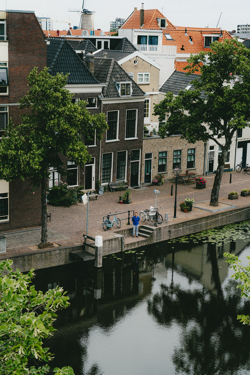 A photo of Dammes who is standing next to a canal. The photo is taken from a distance. In the back, you see a street with trees and some nice old buildings.